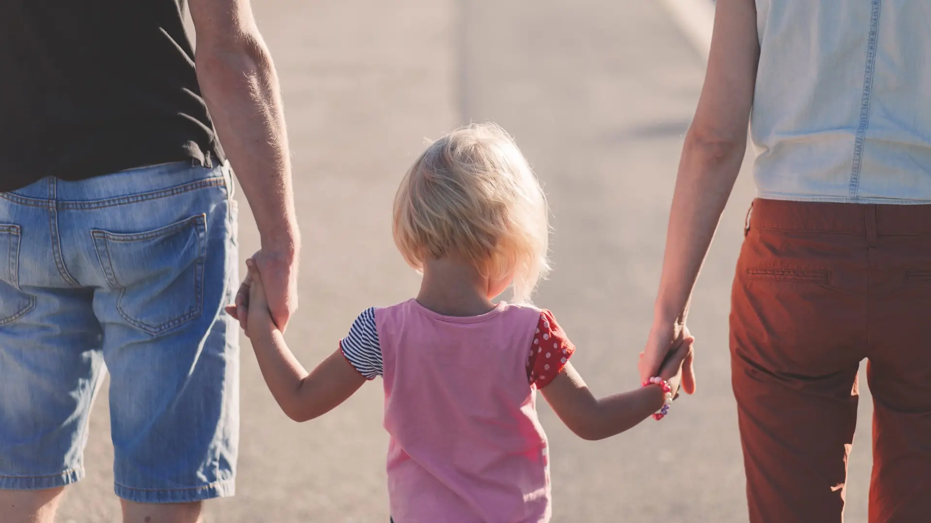A young blonde girl holding hands with her parents from behind.