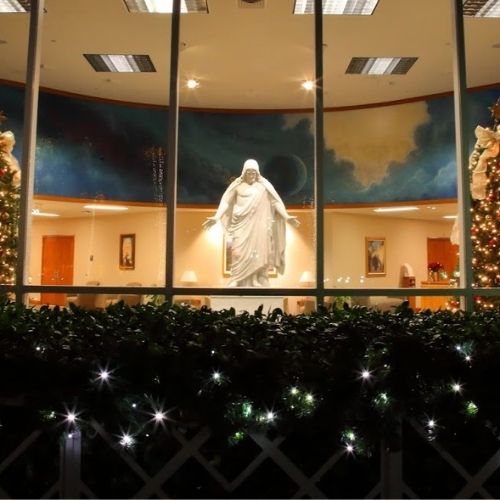 The Christus statue as seen outside the Oakland California Temple Visitors' Center during the Christmas season. Bushes housing faint Christmas lights rest just outside the glass of the Visitors' Center.