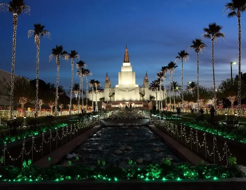 The Oakland California Temple and grounds at Christmas. Christmas lights cover the palm trees and trees, lining the stream on Temple Hill on either side.